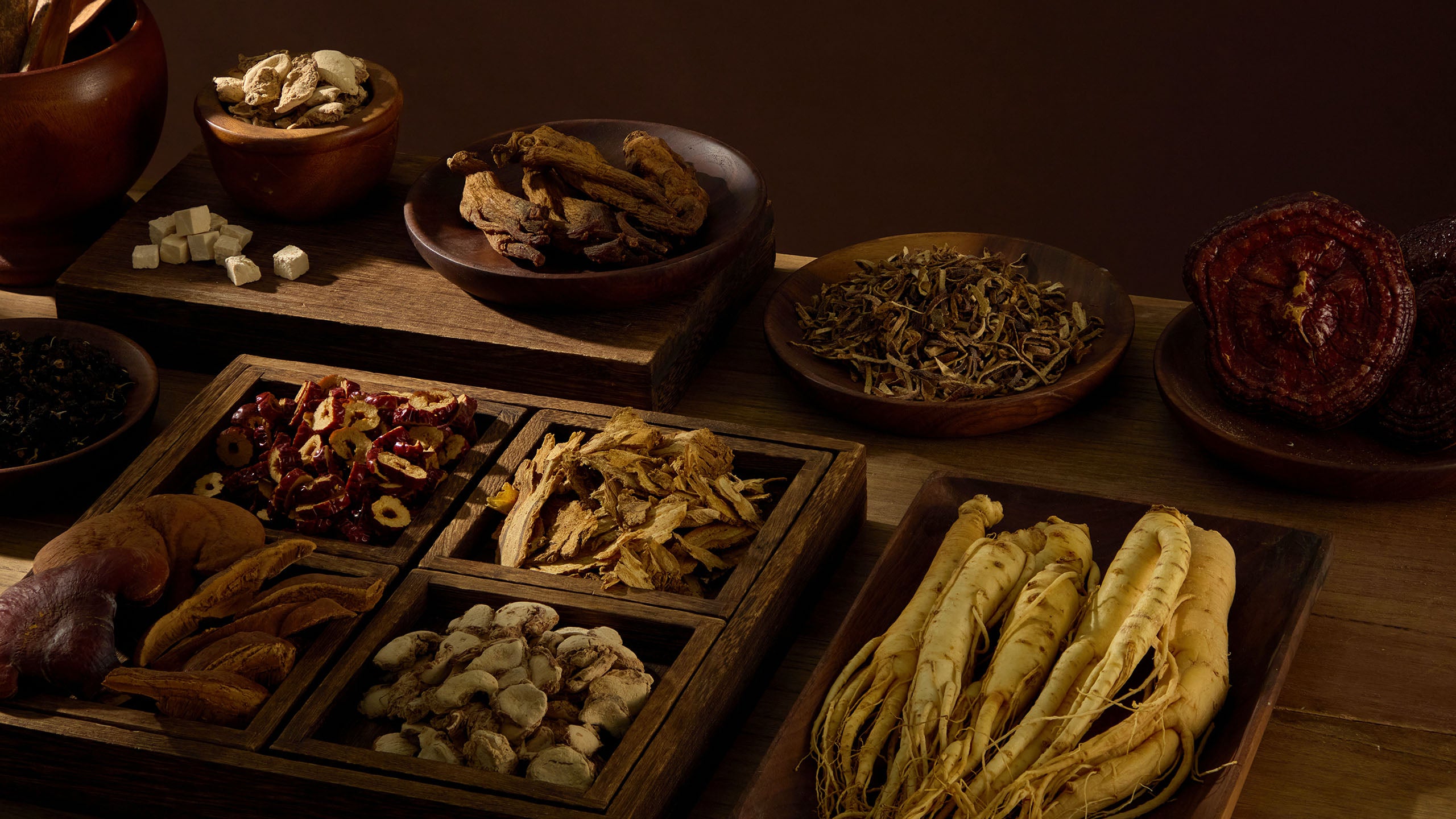 Various dried Chinese herbs and roots on wooden trays and bowls against a dark background.