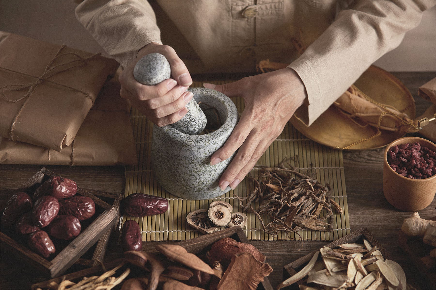 Man using a mortar and pestle with various dried Chinese herbs on a wooden surface.