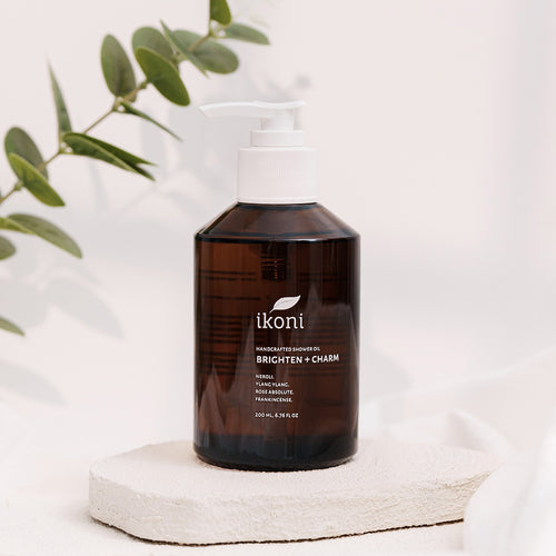 A brown glass bottle with white pump of ikoni Brighten + Charm shower oil against a white background with green leaves.