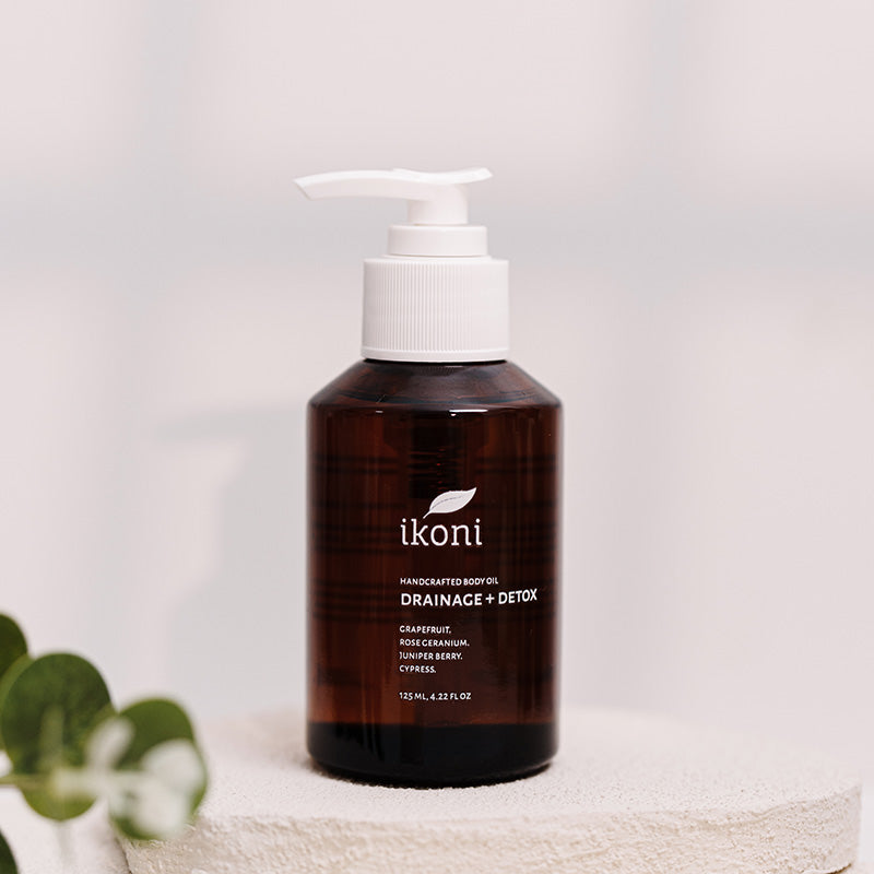 A brown glass bottle with white pump of ikoni Drainage + Detox body oil against an off-white background.