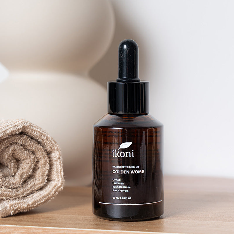 A brown glass bottle with black dropper of ikoni Golden Womb body oil on a wooden surface with a rolled towel in the background.