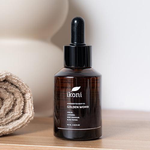 A brown glass bottle with black dropper of ikoni Golden Womb body oil on a wooden surface with a rolled towel in the background.