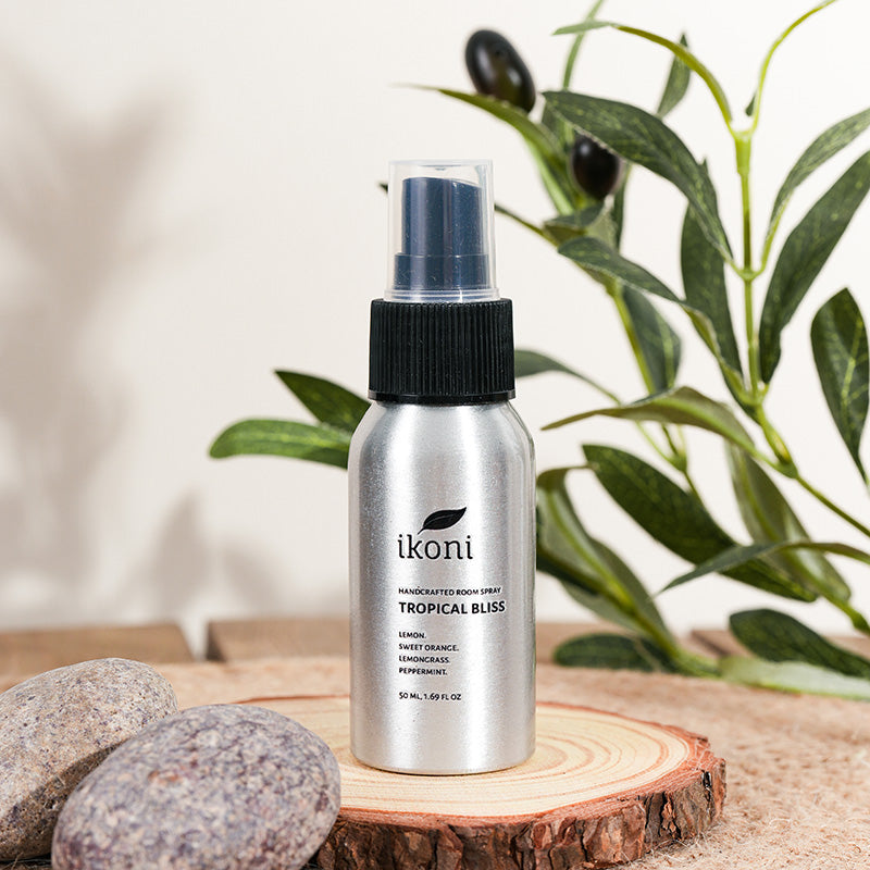 Polished metal spray bottle with black top of ikoni Tropical Bliss on a wooden surface with greenery in the background.