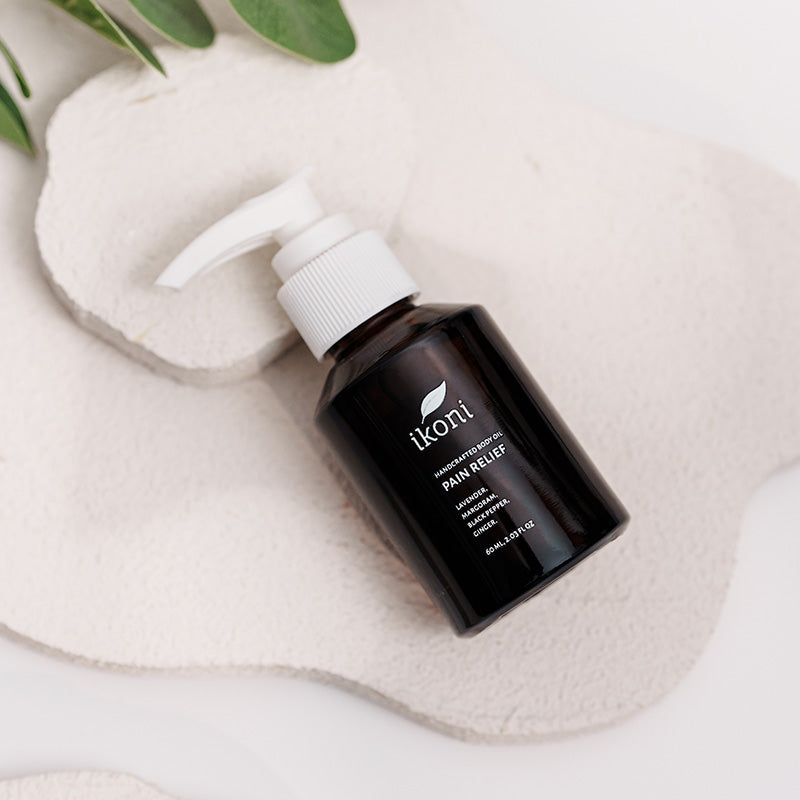 A brown glass bottle with white pump of ikoni Pain Relief body oil on a white stone.
