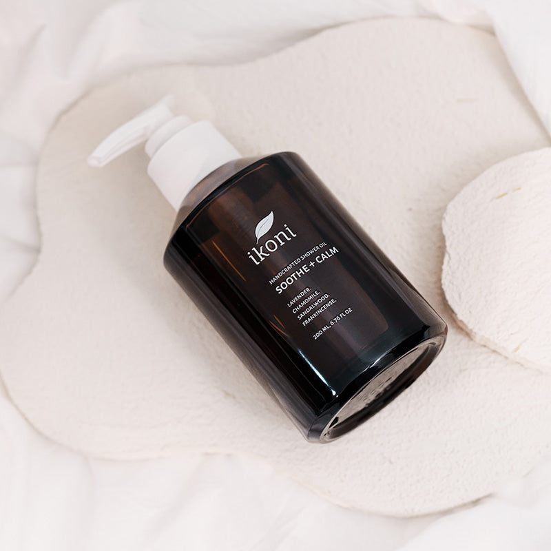 A brown glass bottle with white pump of ikoni Sooth + Calm shower oil on a white stone.