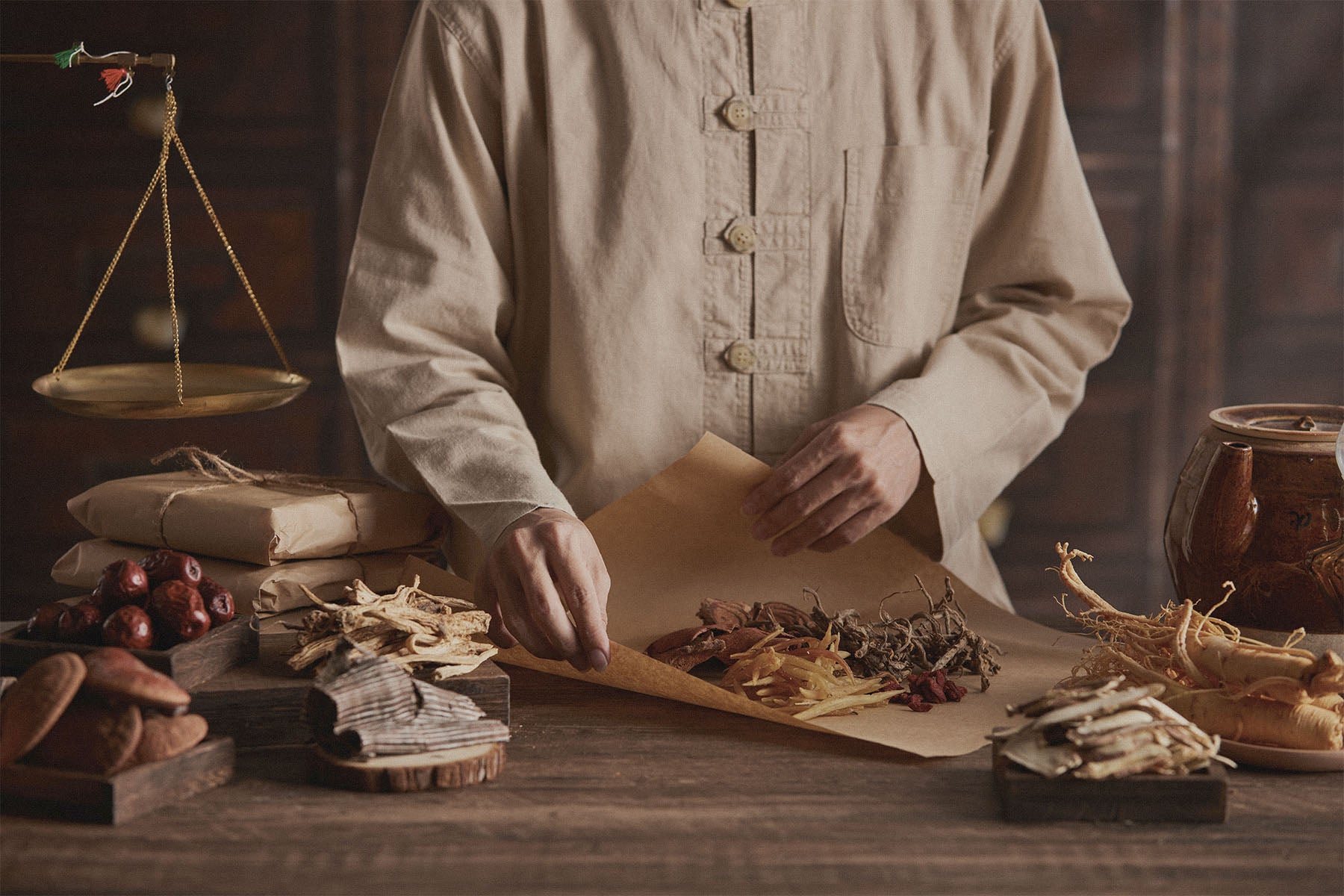 Person in a beige shirt arranging dried Chinese herbs on a table with an antique scale and other items in the background.